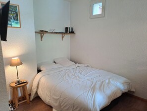 A bedroom with a single bed, a bedside table with a lamp, a small shelf above the bed, and a window.
