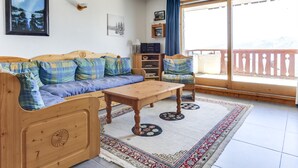 A cozy living room with a wooden sofa, a wooden coffee table, and a chair. There is a large window with a view of mountains. - Charming 4-room duplex, 8 people, west-facing terrace, near slopes (Peisey-Vallandry)