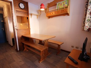 A wooden dining table with benches, a wall clock, a shelf with children's drawings, and a television on a side table.