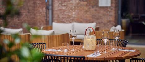 An outdoor dining area with a brick wall in the background, wooden tables, and black chairs.