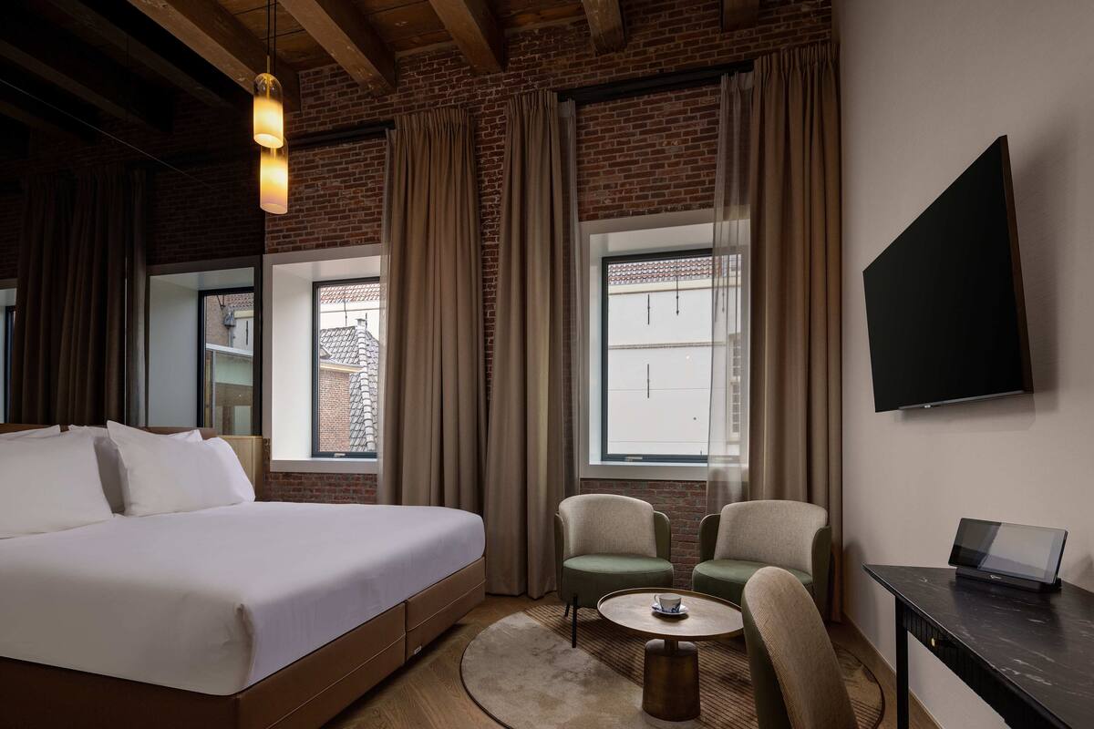 Habitación de hotel con una cama grande, dos sillones, una mesita, televisión y ventana con cortinas.