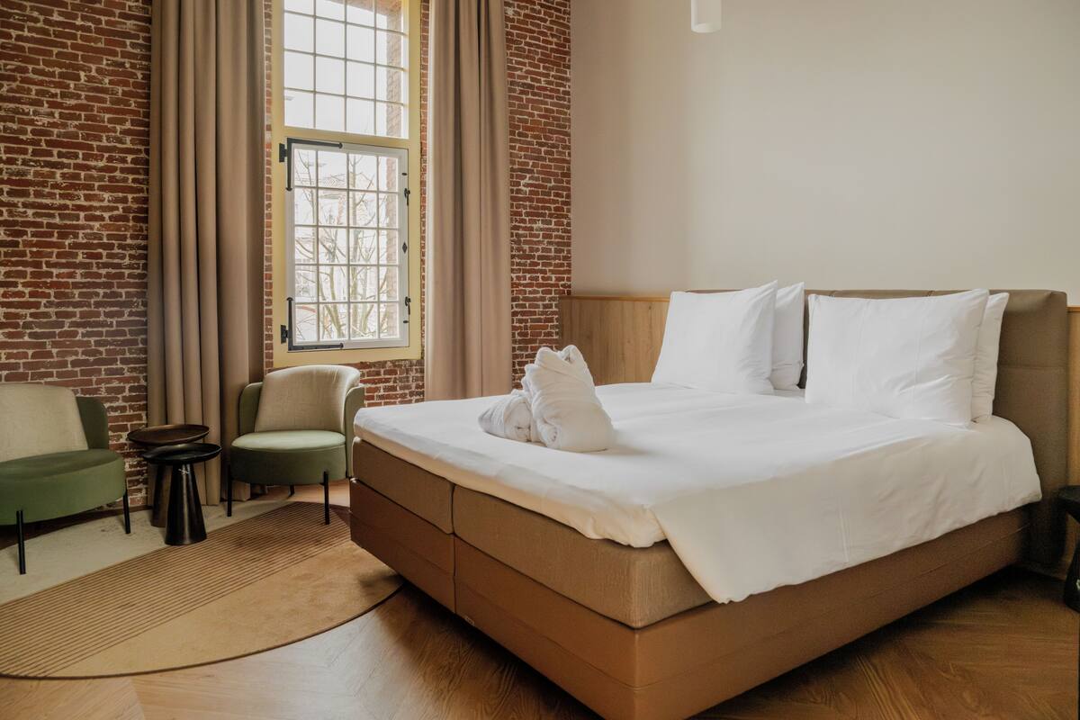 Habitación de hotel con pared de ladrillo, un ventanal con cortinas, cama con ropa blanca, un sillón verde y una mesita redonda.