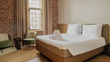 Een hotelkamer met een bakstenen muur, een groot raam met gordijnen, een bed met witte lakens, een groene fauteuil en een kleine ronde tafel.
