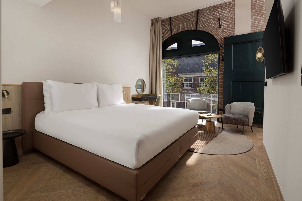 Una habitación de hotel moderna con una cama grande, una pequeña mesa redonda, una silla y vista a un edificio de ladrillo a través de una ventana en arco.