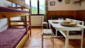 Un lit superposé avec une couvre-lit à motifs rouges et verts, une petite table avec une assiette et des verres, une chaise, et une vue sur l’extérieur par une fenêtre.