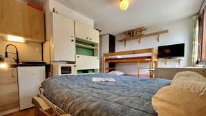 Une chambre de taille modeste équipée de lits superposés, d’un coin cuisine, d’une télévision et d’un petit bureau.
