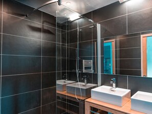 Een moderne badkamer met donkere betegelde wanden, een douchecabine met glazen wanden en twee witte wasbakken op een houten wastafelmeubel.