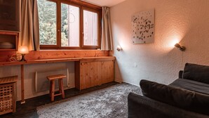 A cozy room with a wooden desk, a window with curtains, a radiator, a small stool, a dark sofa, and a wall-mounted light fixture. - Cozy studio for 2, near slopes and shops with Wifi, parking Arc 1800 (Les Arcs 1800)