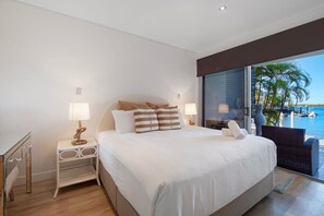 A bedroom with a large bed, two bedside tables with lamps, a view of the sea through a glass door, and a wicker chair by the door. - Waterfront Oasis in Runaway Bay (Runaway Bay)