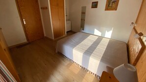 Ein Schlafzimmer mit Holzboden, einem Bett mit weißer Bettdecke, einem Holzschrank, einem Nachttisch mit Lampe und einem kleinen Bild an der Wand.