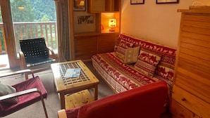 Ein gemütliches Zimmer mit einer Holzcouch, einem kleinen Tisch und einem Sessel. Ein Fenster bietet Blick auf Bäume.