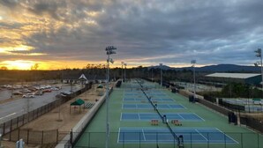 A tennis court complex with multiple courts, seating areas, and a parking lot.
