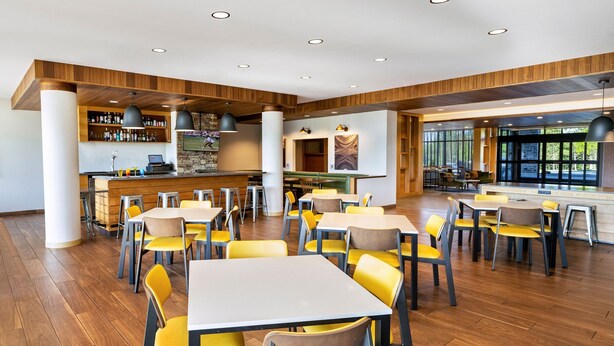 A modern restaurant with wooden flooring, yellow and brown chairs, and a bar area with a stone wall and shelves of bottles.
