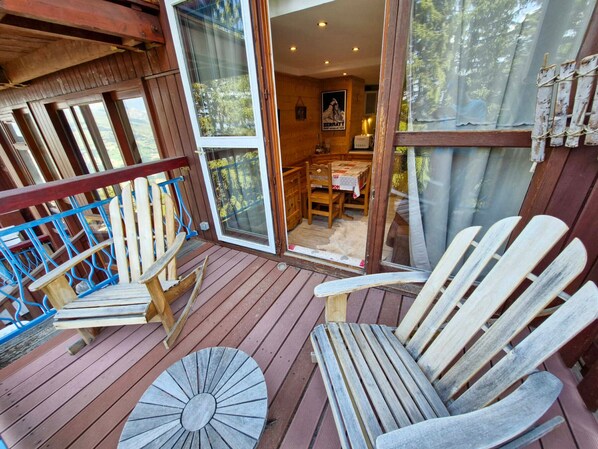 A wooden deck with Adirondack chairs, a table, and a glass door leading to an interior dining area.