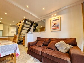 A living room with a brown sofa, a dining table with a white tablecloth, and a staircase with a wooden railing.