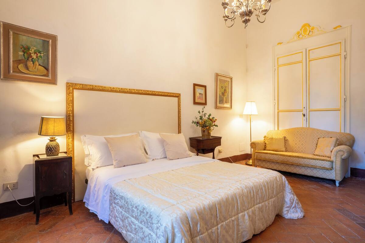 A bedroom with a large bed, a sofa, a nightstand with a lamp, and a wall with framed artwork.