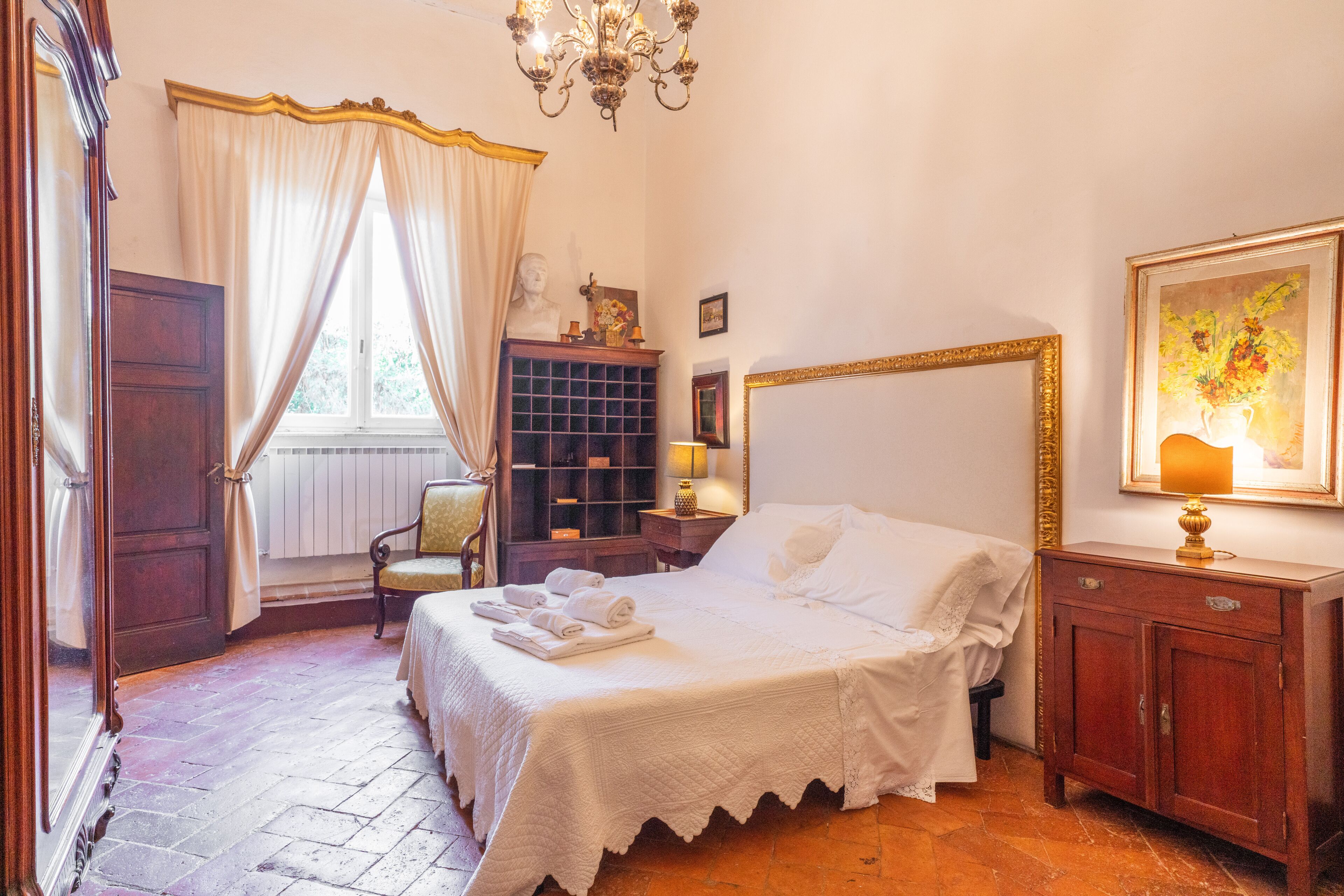 Une chambre à coucher avec un grand lit, une table de chevet en bois, une commode, une chaise, une bibliothèque et un lustre.