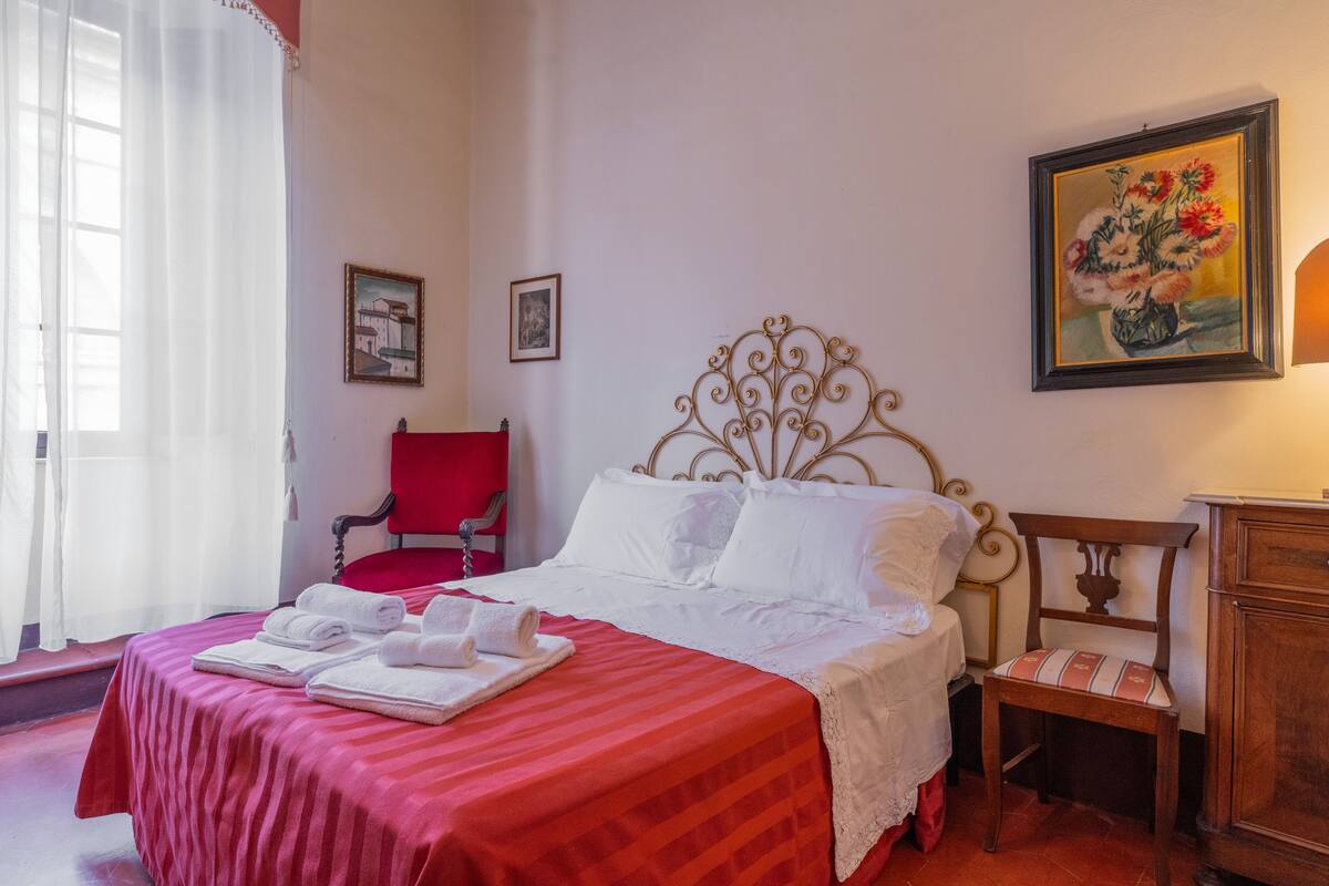 A neatly made bed with white linens and red bedding, a red chair, a wooden nightstand, and a painting on the wall.