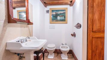 A bathroom with a sink, toilet, and bidet, wooden beams on the ceiling, and a painting on the wall.