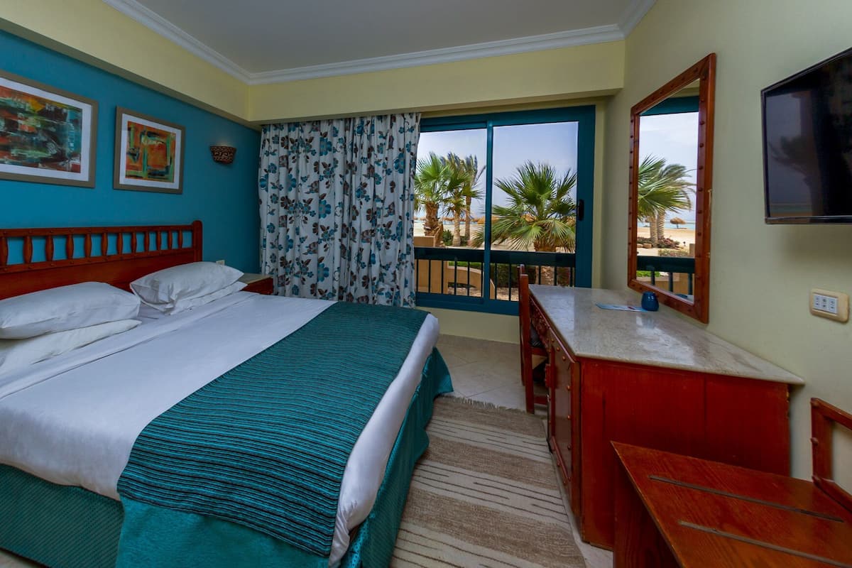 Ein Hotelzimmer mit Bett, Schreibtisch, Fernseher und einem Balkon mit Blick auf Palmen und den Strand.