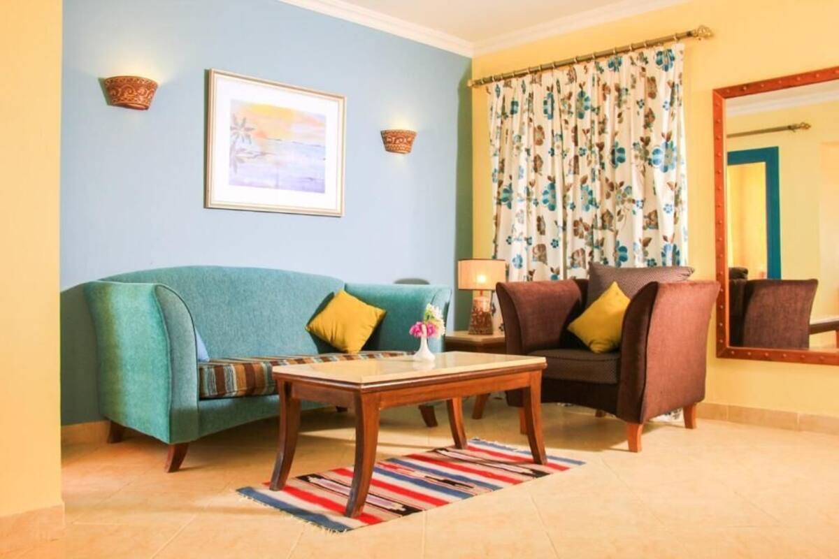 Ein Wohnzimmer mit einem blaugrünen Sofa, einem braunen Sessel, einem Holztisch und einem farbenfrohen StreifenTeppich.