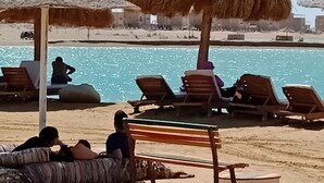 Ein Strand mit Liegestühlen, einem Pool und Reetdachschirmen.
