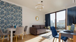A modern living room with a sofa, dining table, and chairs. There is a laptop on the table, and a large window with curtains.