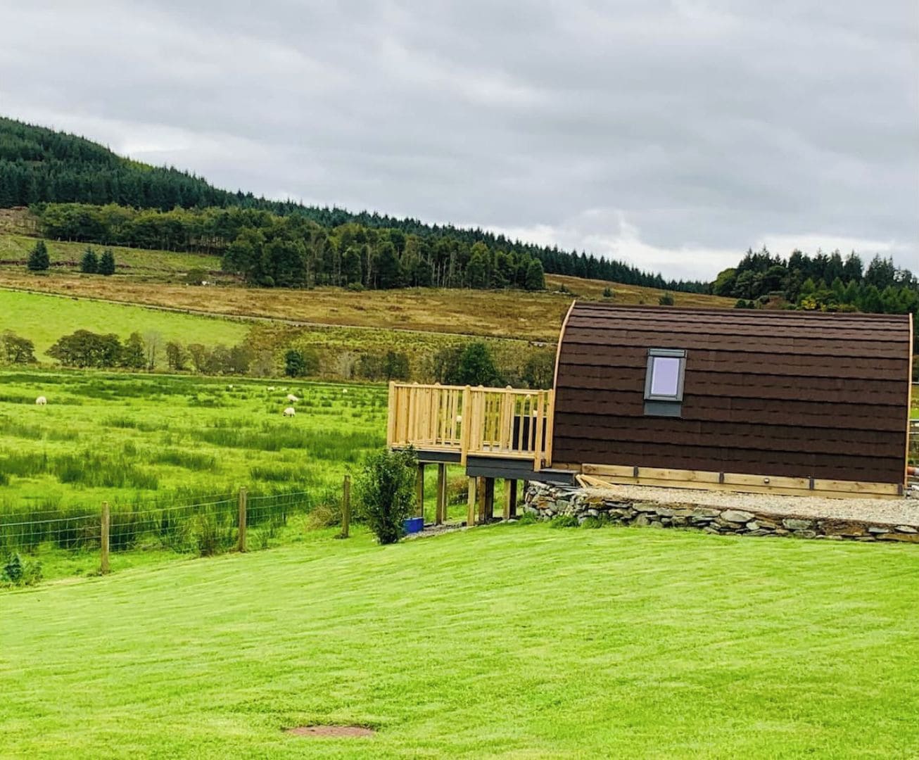 Un chalet rustique doté d’une terrasse en bois, entouré de prairies verdoyantes et bordé, au loin, par une colline boisée.