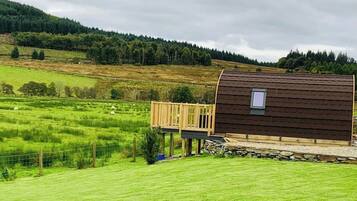 Un chalet rustique doté d’une terrasse en bois, entouré de prairies verdoyantes et bordé, au loin, par une colline boisée.