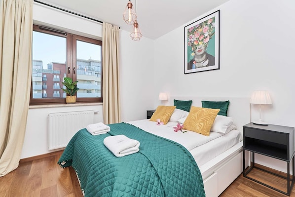 A bedroom with a bed, a window with curtains, a radiator, a bedside table, and a wall-mounted picture.