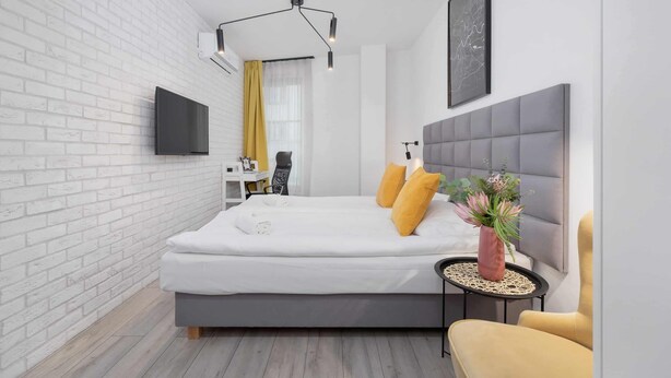 Ein modernes Schlafzimmer mit weißer Ziegelwand, einem grauen Bett, einem angebrachten Fernseher, einem Nachttisch mit einer Vase und einem gelben Sessel.
