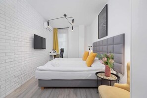 Quarto moderno com parede de tijolos brancos, cama cinza, TV montada, mesa de cabeceira com um vaso e uma cadeira amarela.