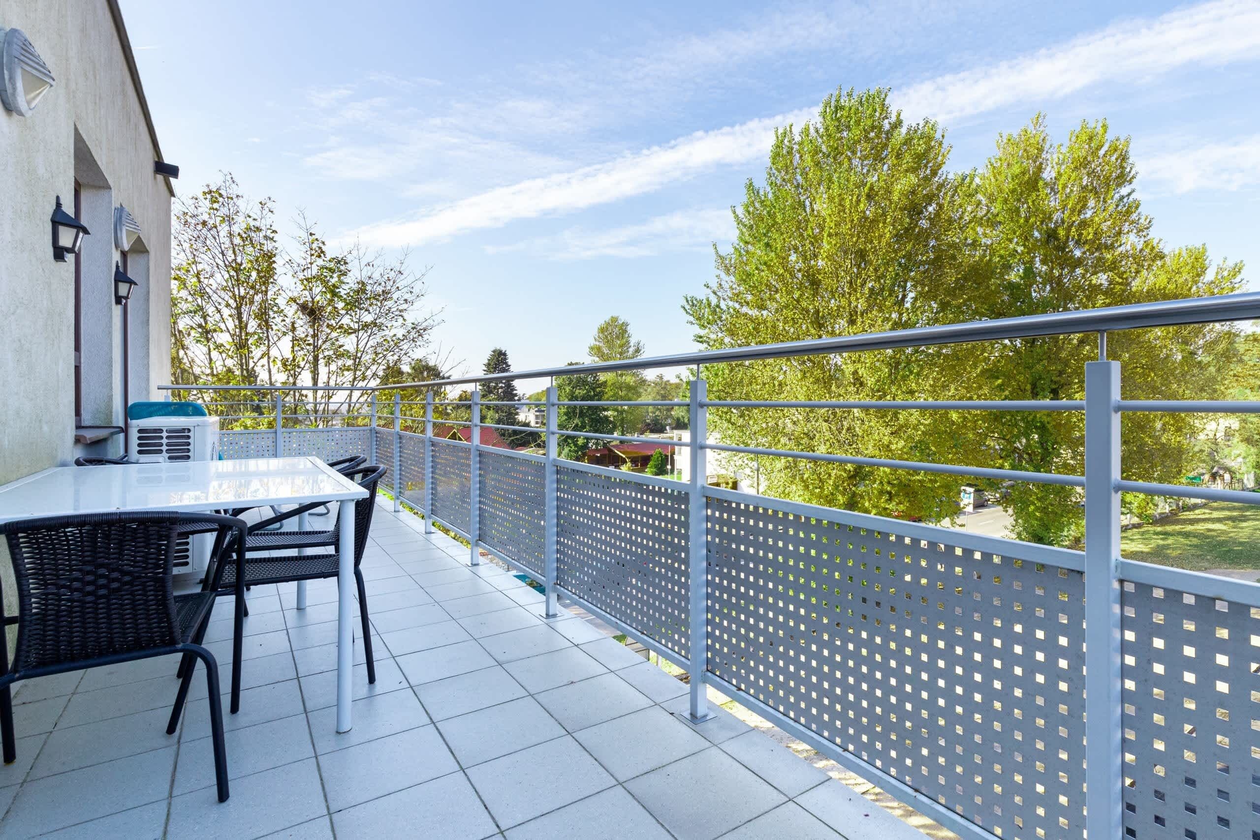 Une terrasse extérieure avec une table et des chaises, une rambarde en métal et des arbres en arrière-plan.