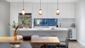 A modern kitchen with a central island, pendant lights, a large window with a forest view, and a dining area with a wooden table and chairs. - Kangary- secluded bush retreat with spa (Daylesford)