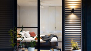 A modern bedroom with a bed, pillows, and a bedside table. There is a window with blinds, a small plant, and a wall-mounted light fixture. - Kangary- secluded bush retreat with spa (Daylesford)