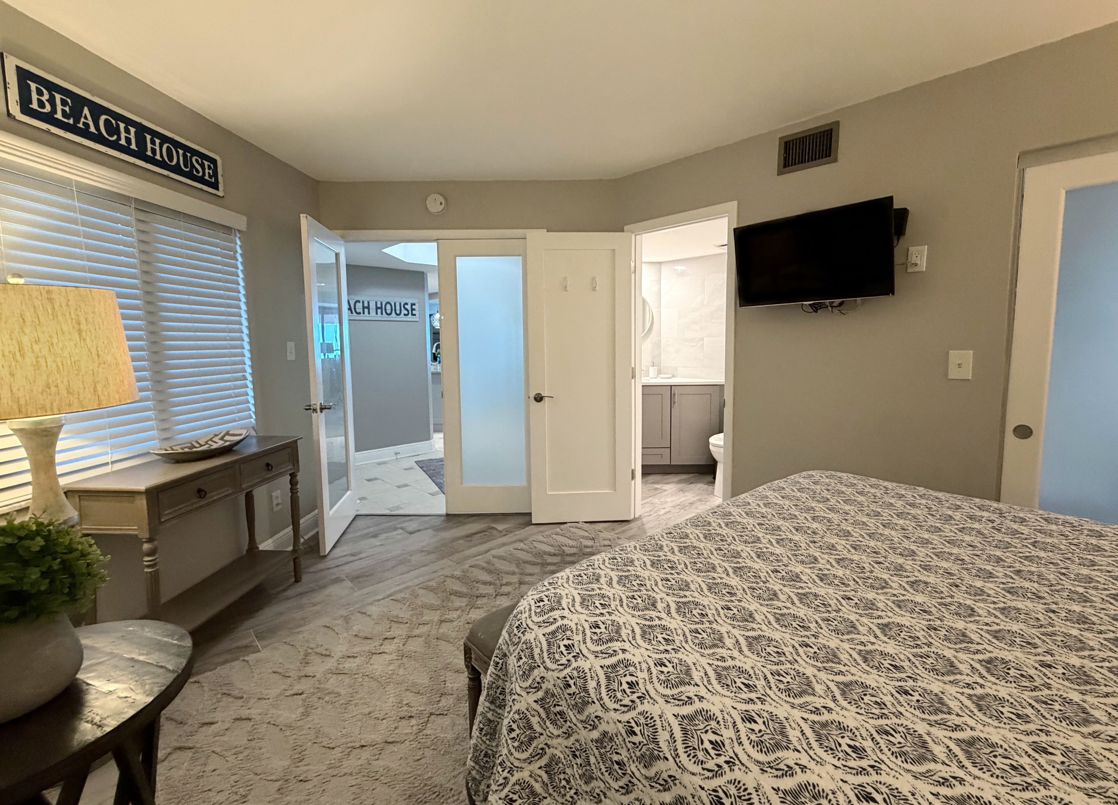 A bedroom with a bed, a television, a desk, and a chair. There is a hallway leading to a bathroom and a kitchen.