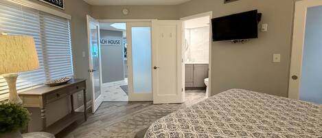 A bedroom with a bed, a television, a desk, and a chair. There is a hallway leading to a bathroom and a kitchen.