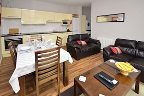 Una sala moderna con comedor, una cocina al fondo y un sofá de cuero negro con un frutero sobre una mesa de centro.