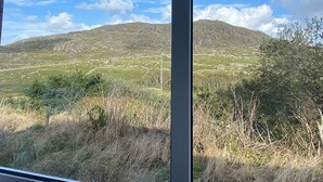 Blick auf eine gebirgige Landschaft durch ein Fenster, mit einer grünen Wiese und blauem Himmel.