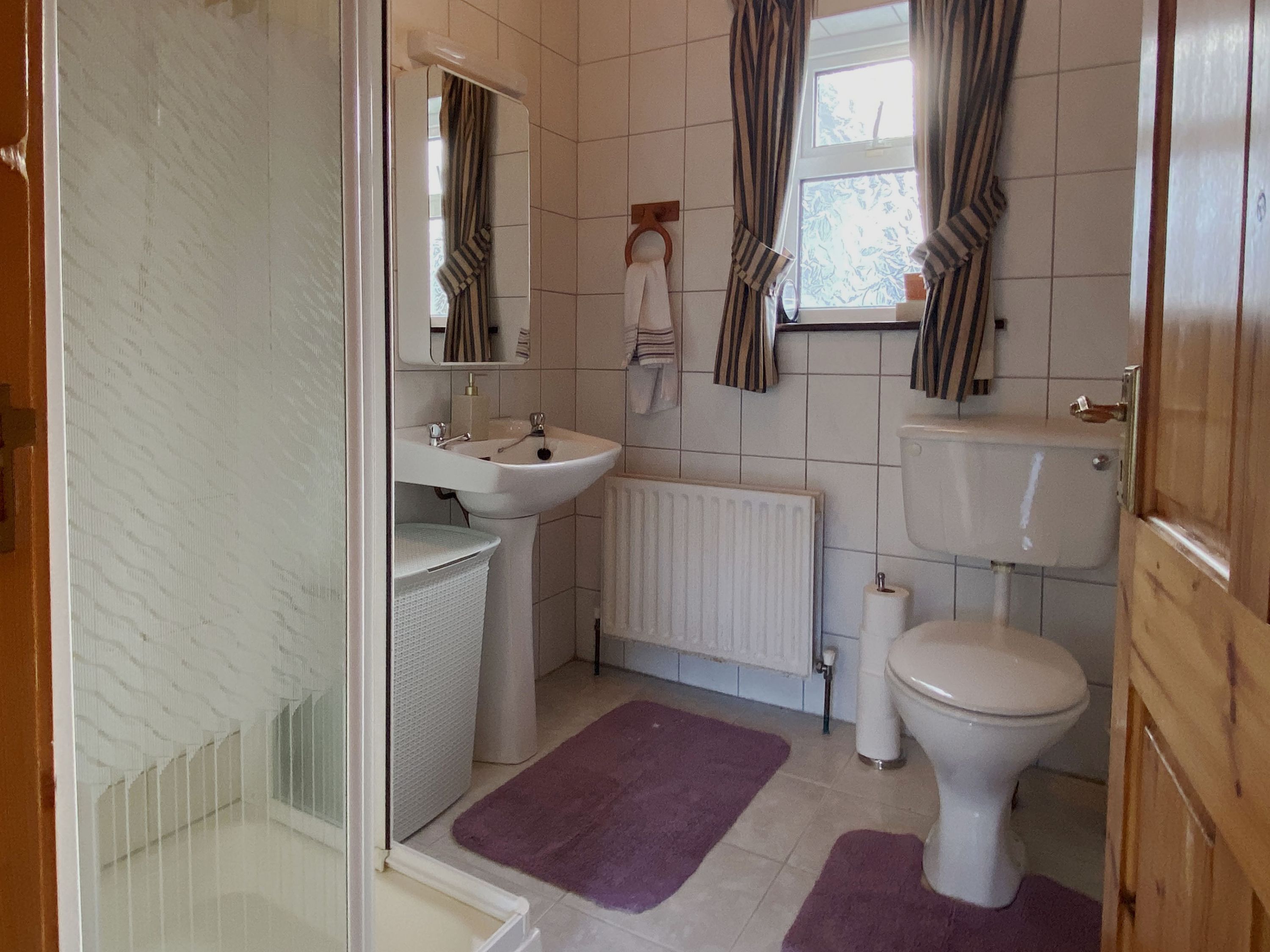Ein Badezimmer mit einem weißen Waschbecken, einer Toilette und einem Heizkörper. An den Fenstern hängen gestreifte Vorhänge und auf dem Boden liegt ein violetter Teppich.