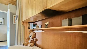 A small, cozy room with a wooden headboard, a bed with a rolled towel, a teddy bear, and a shelf with books and a small vase.
