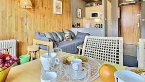 A cozy room with a wooden ceiling, a grey sofa with cushions, a wicker chair, and a dining table with cups and a fruit bowl.