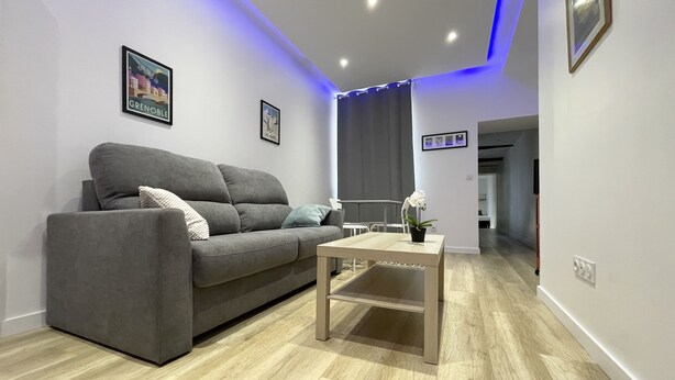 Uma sala de estar moderna com um sofá cinzento, uma mesa de centro de madeira e uma orquídea em vaso. Há quadros nas paredes e uma escadaria ao fundo.