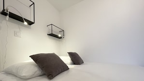 Uma cama bem arrumada com roupa de cama branca e duas almofadas escuras, um candeeiro fixado na parede e uma tomada do lado esquerdo da cama.