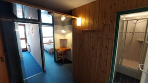A narrow hallway with a wooden wall, a desk with a chair, a bed in the background, and a shower area.