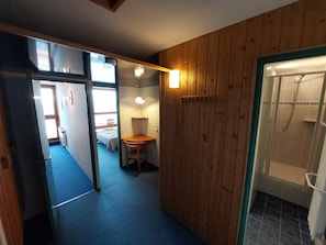 A narrow hallway with a wooden wall, a desk with a chair, a bed in the background, and a shower area.