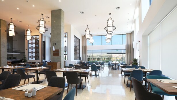 A modern dining area with multiple tables and chairs, large windows, and hanging pendant lights. - Park Dedeman Adıyaman (Adiyaman)