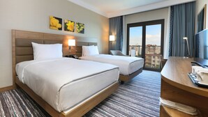 A hotel room with two beds, a desk with a lamp, a TV, and a view of the city. - Park Dedeman Adıyaman (Adiyaman)