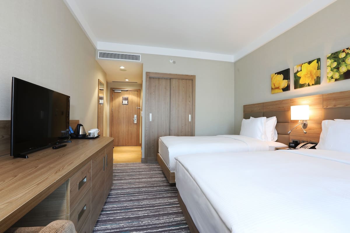 A hotel room with a TV, wooden desk, two beds, a nightstand, and a wall-mounted lamp.