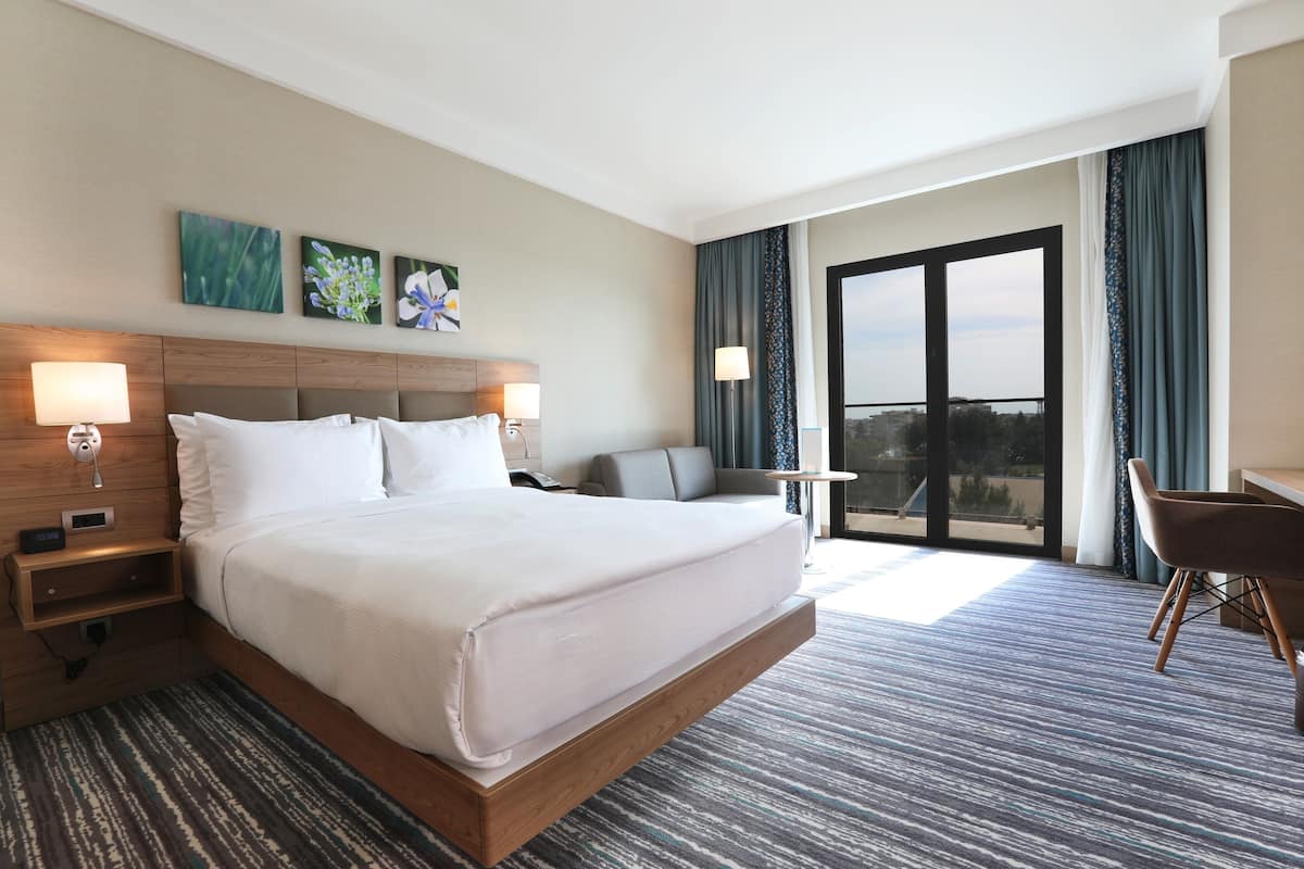 A modern hotel room with a large bed, bedside tables, a desk, and a view of the city through the balcony.